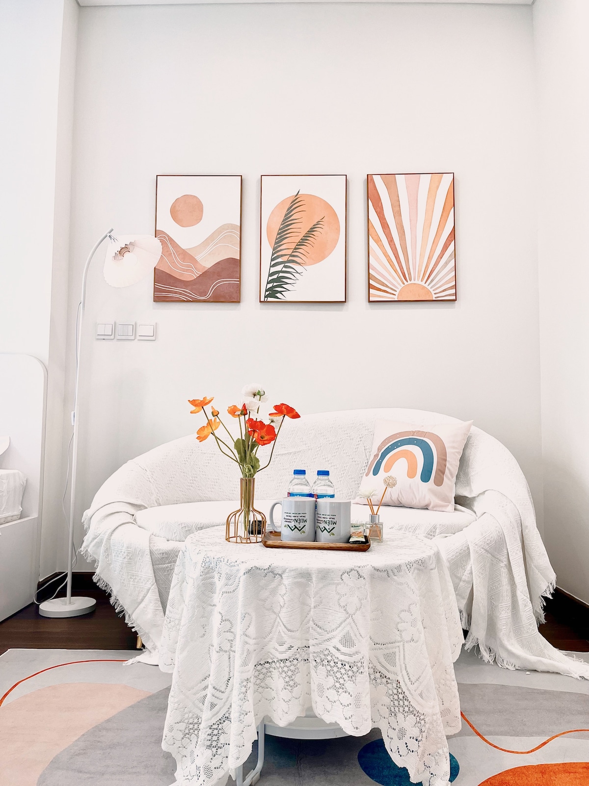 A cozy seating area features a round white couch adorned with a light throw. A small table holds decorative items, including a vase with flowers and two bottles of water. Colorful artwork hangs on the wall, complemented by a patterned rug beneath.