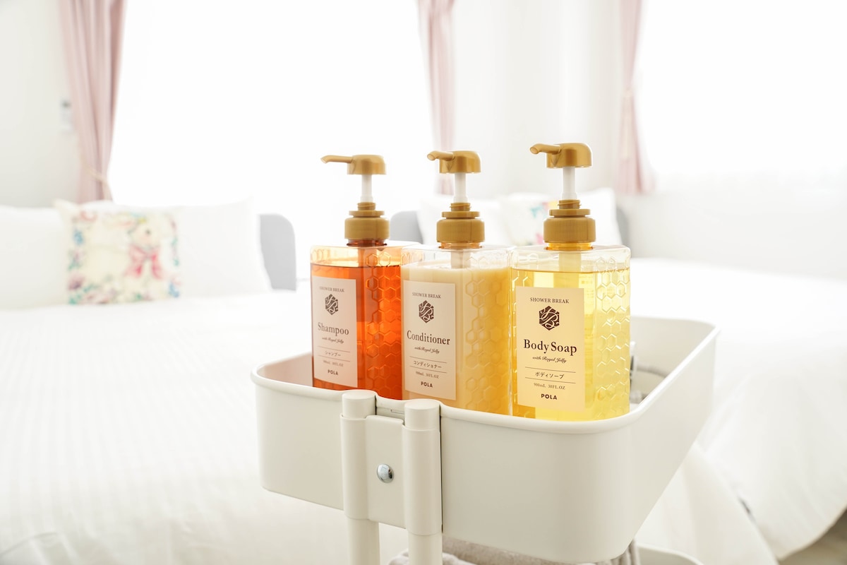 A set of three pump bottles is displayed on a small cart, containing shampoo, conditioner, and body soap. Natural light streams through nearby windows, illuminating the soft color palette of the room, which features a spacious bed in the background adorned with decorative pillows.