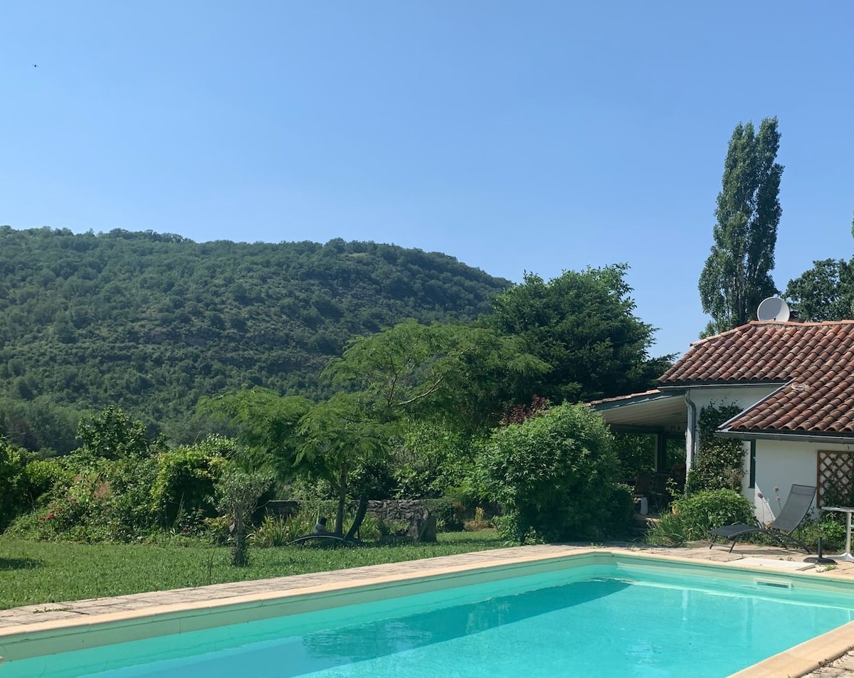 The image showcases a serene outdoor scene featuring a swimming pool surrounded by lush greenery. Rolling hills can be seen in the background under a clear blue sky, creating a peaceful atmosphere. The pool area is equipped with sun loungers, inviting relaxation.