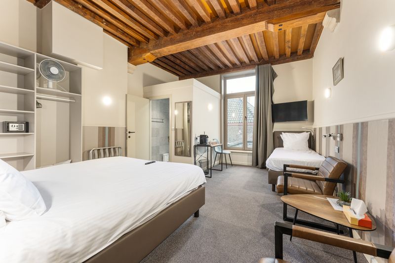 A cozy triple room features a spacious double bed and a separate single bed. Exposed wooden beams enhance the room's character, while natural light enters through a window. A smart TV is mounted on the wall, and a small seating area with a table is included.