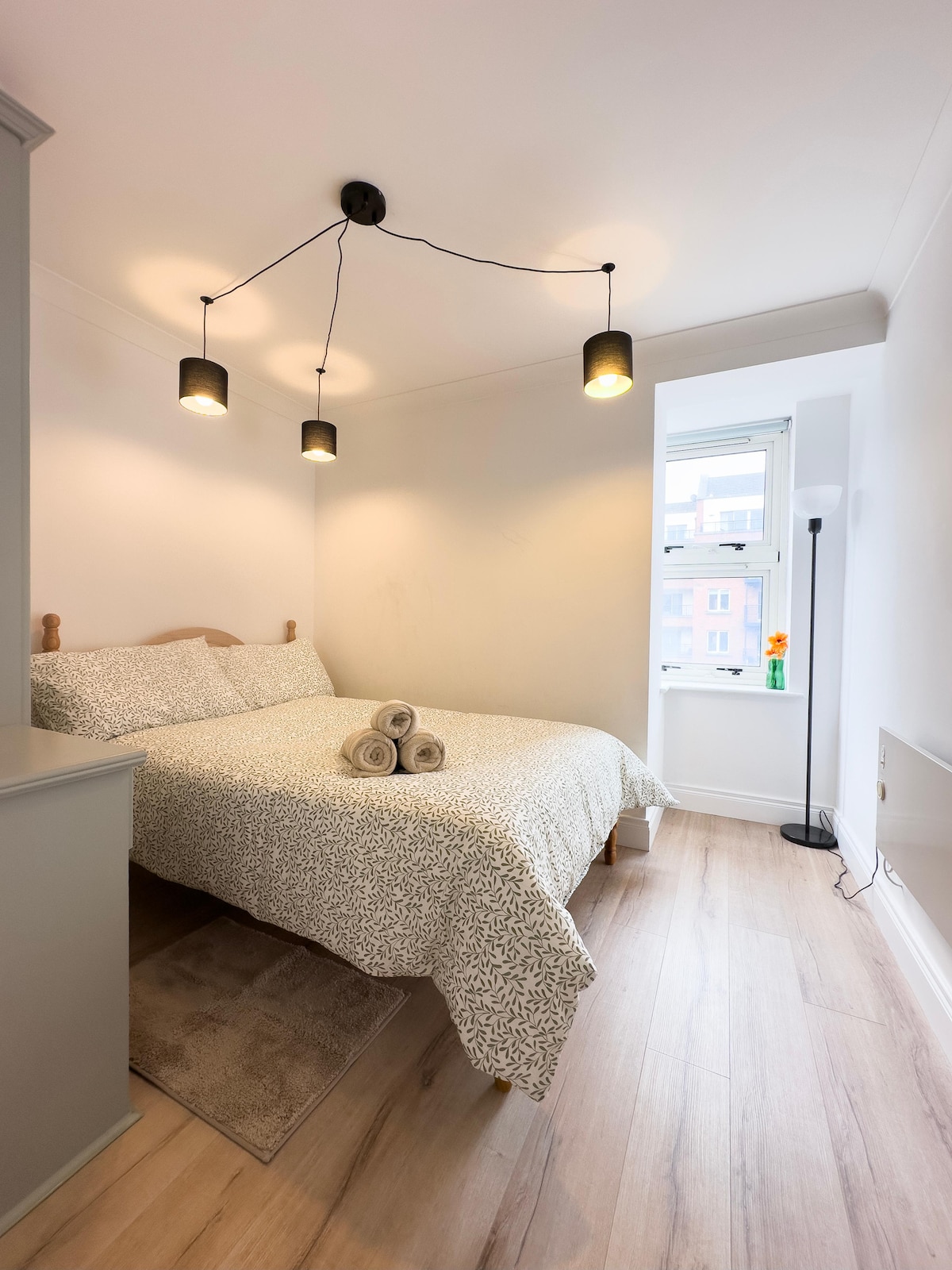 A cozy double room is featured, with a spacious bed adorned with a patterned duvet. Three pendant lights illuminate the area above the bed. A small window provides natural light, and neatly rolled towels are arranged on the bed. Light wooden flooring enhances the room's brightness.
