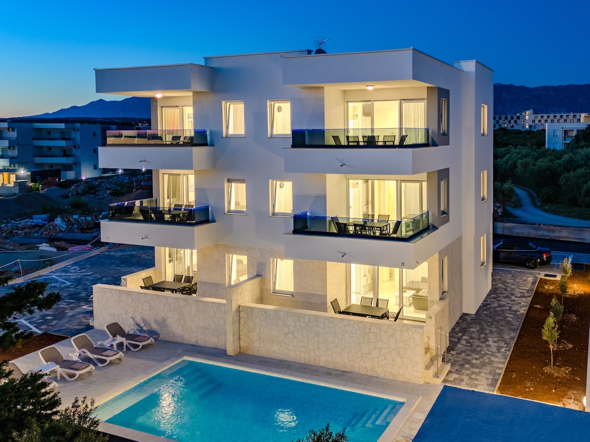 A modern three-story building is illuminated at dusk, featuring balconies with sliding glass doors. A well-maintained swimming pool is situated in front, surrounded by sun loungers. The property is landscaped with greenery, providing a calm and relaxing environment.