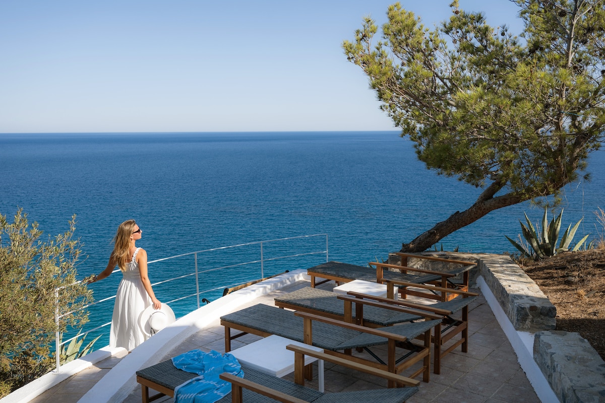 An outdoor terrace overlooks the serene sea, furnished with wooden chairs and a table. A gentle slope of greenery leads to the water, creating a peaceful atmosphere. The clear sky complements the tranquil view, enhancing the appeal of this private space.