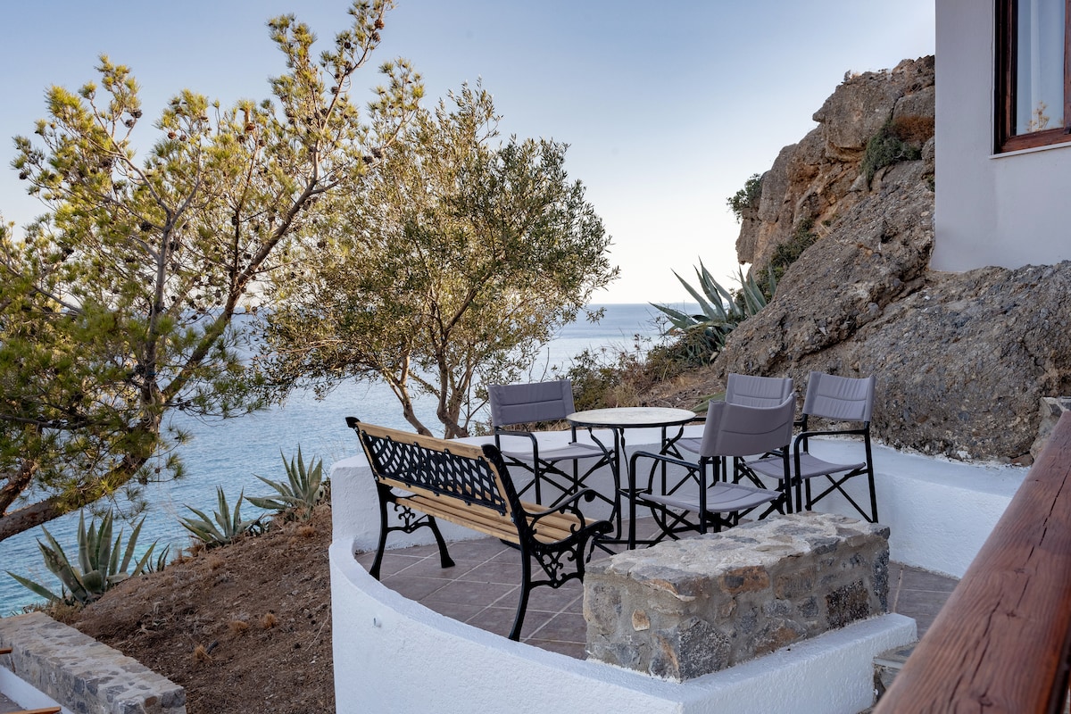 An outdoor seating area is presented, featuring a round table surrounded by four chairs. Rock formations and a few trees can be seen nearby, alongside glimpses of the sea in the background, creating a relaxed atmosphere for outdoor enjoyment.