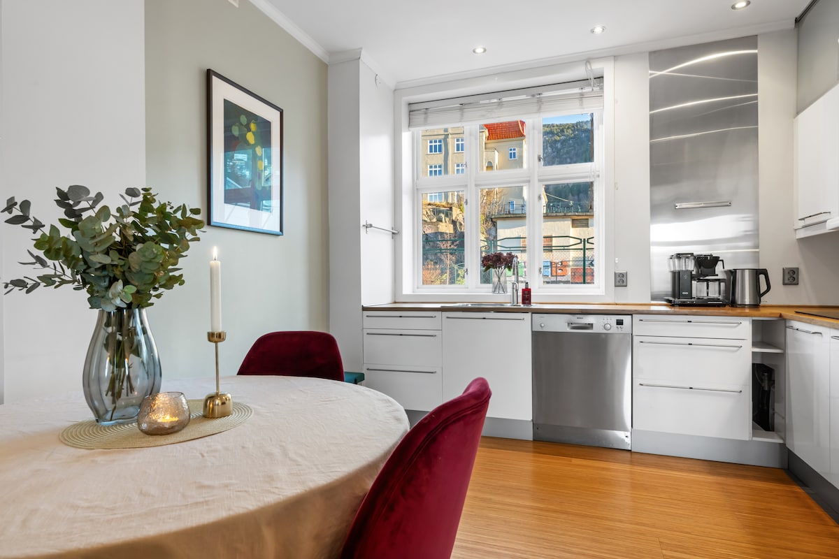 A modern kitchen area is highlighted by sleek cabinetry and stainless steel appliances, including a dishwasher and coffee machine. A round dining table is set with an elegant tablecloth and a small vase of greenery. A large window provides natural light and a view of the exterior architecture.