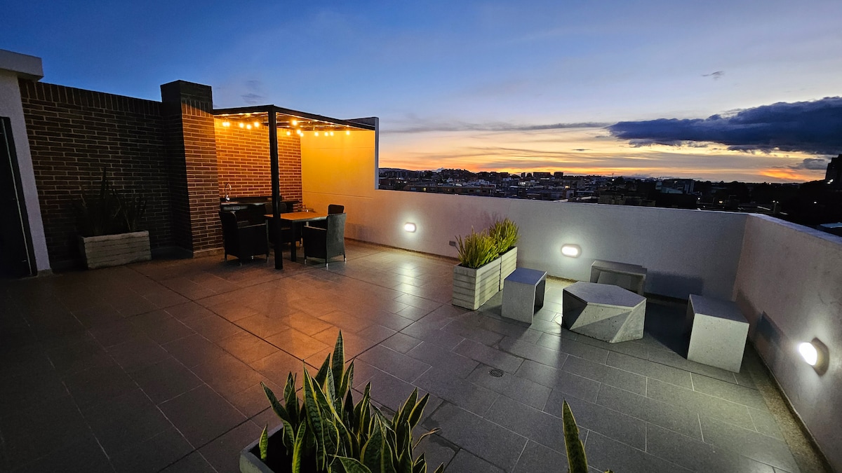 A rooftop terrace is illuminated by subtle lighting as day transitions to night. A dining area with dark furniture is situated under a covered pavilion. Planters with greenery are placed around the space, complemented by modern stone seating and a panoramic view of the skyline.