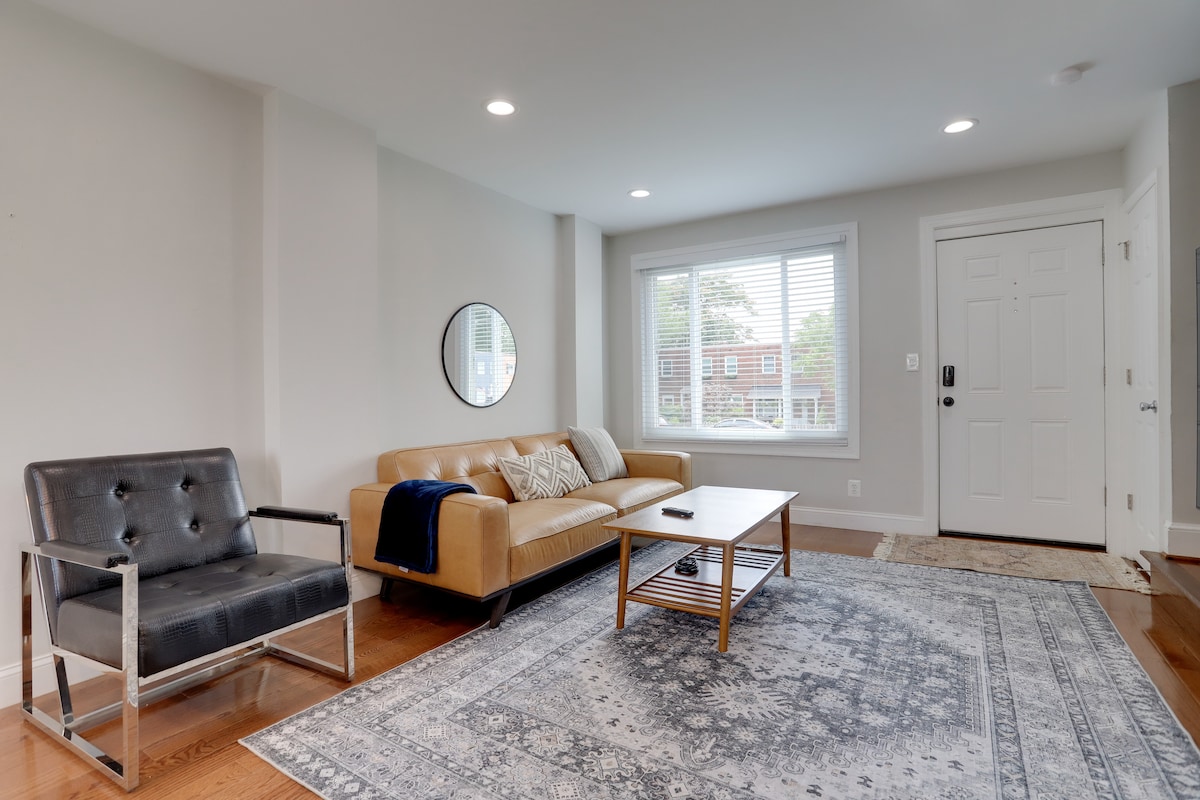 A comfortable living area is presented with a stylish gray armchair and a tan sofa. A wooden coffee table is centered on a decorative area rug. Natural light fills the space through the window with blinds, highlighting the neutral color palette and inviting ambiance.