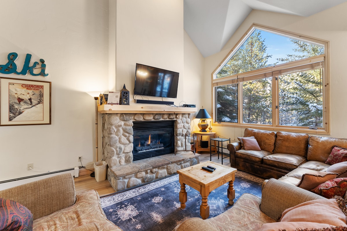 A cozy living area features a large sectional sofa and a pair of comfortable chairs arranged around a stone fireplace. A flat screen TV is mounted above the fireplace, with natural light streaming through expansive windows that provide views of the surrounding trees.