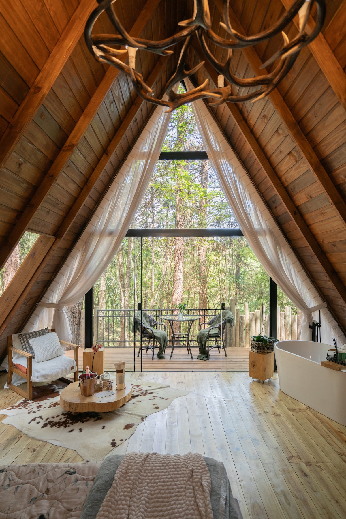 The interior features high wooden ceilings with a modern chandelier, allowing natural light to fill the space through large glass windows. A cozy seating area includes a soft chair and circular coffee table. An inviting freestanding bathtub is positioned nearby, enhancing the relaxing atmosphere.