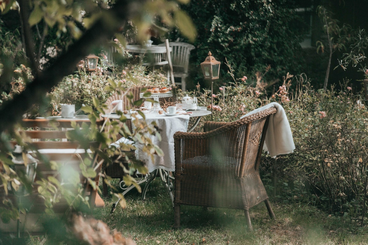 A cozy outdoor seating area is surrounded by lush greenery. A wicker chair is positioned near a table set with tea and pastries, accompanied by white tableware. Soft textures and natural elements create a serene atmosphere, inviting guests to relax in the garden.