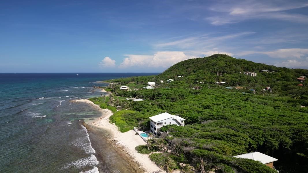 An aerial view highlights the villa nestled along the shoreline of Pumpkin Hill, surrounded by lush greenery. The turquoise waters of the Caribbean gently lapping at the sandy beach create a serene coastal scene. Rolling hills frame the backdrop, enhancing the natural beauty of the location.