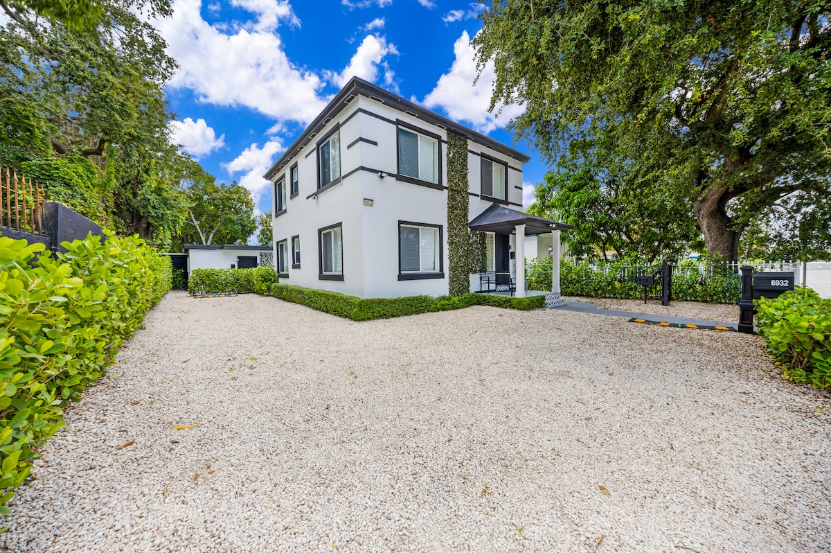 The exterior of a modern two-story apartment building is shown, surrounded by manicured hedges and a gravel driveway. Large windows provide natural light, while greenery frames the property. A covered entrance indicates access to the apartment, enhancing the welcoming environment.