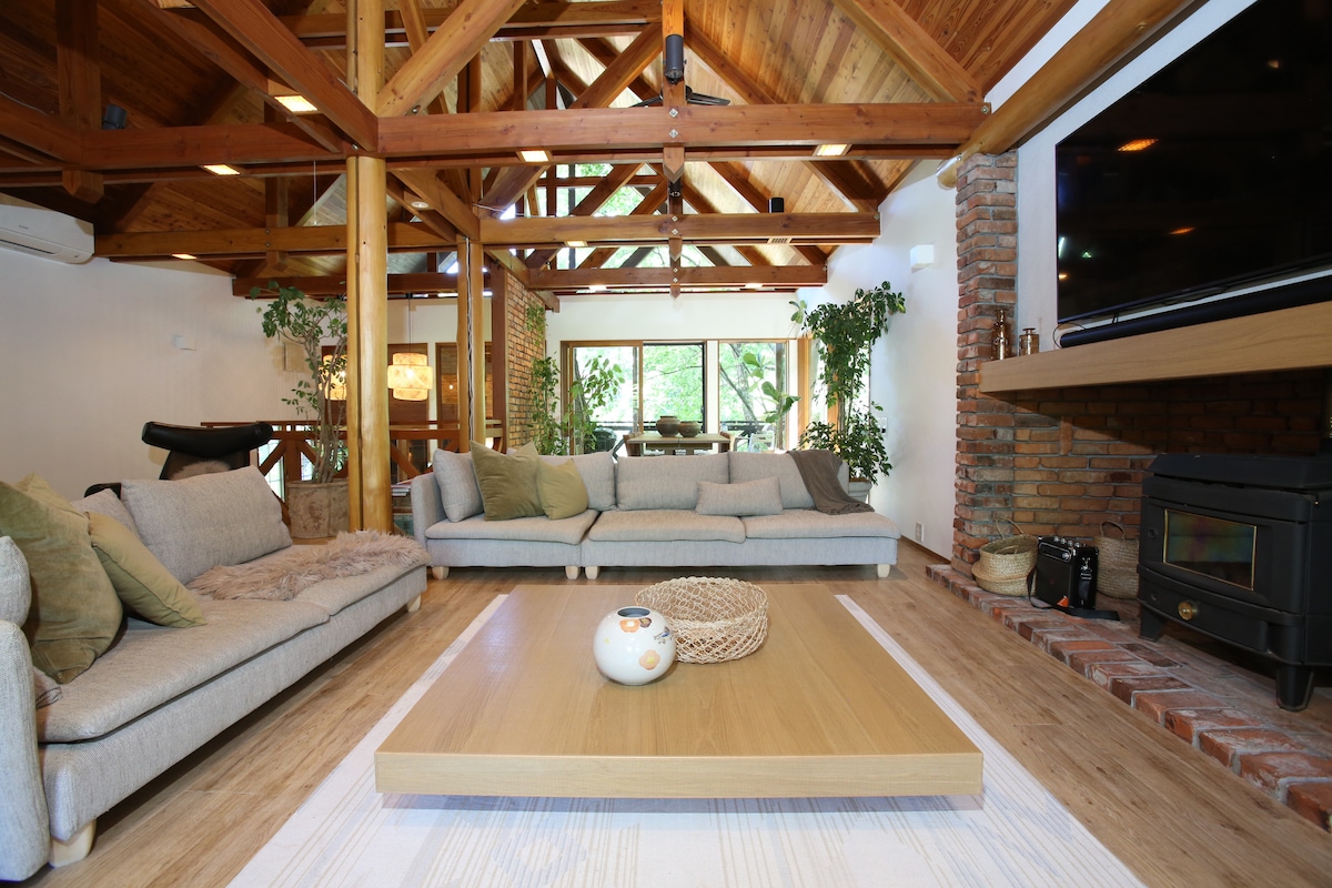 A spacious living room features exposed wooden beams and high ceilings, with natural light filtering through large windows. Comfortable seating areas include soft sofas arranged around a central table. Lush greenery adds a vibrant touch, enhancing the welcoming and relaxed ambiance.
