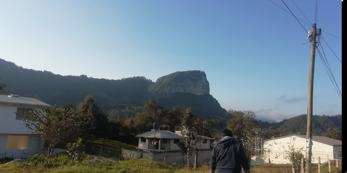 A scenic view captures the prominent Cabezón mountain in the background, framed by trees and greenery. Two buildings, displaying a modern architectural style, are positioned in the foreground, with a clear blue sky above. Power lines are visible along the side.