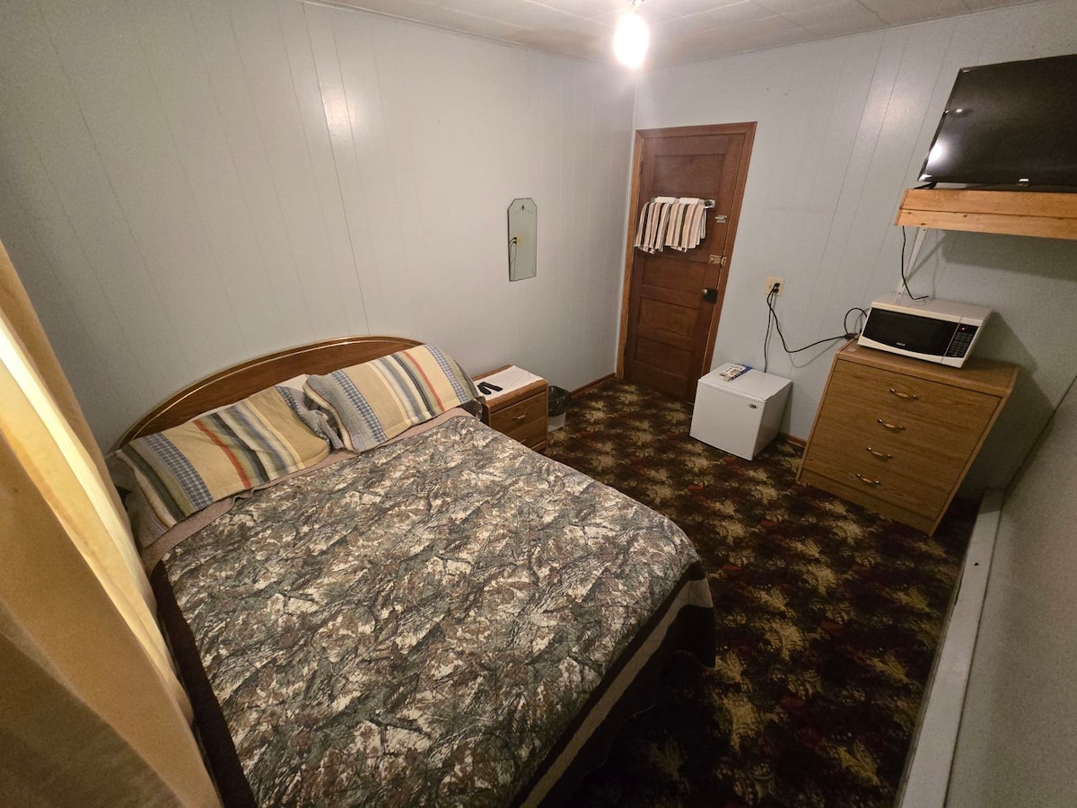 A cozy room features a spacious bed adorned with a camo-patterned blanket, complemented by striped pillows. A small bedside table and a wooden dresser provide additional storage. A microwave and a compact refrigerator are neatly positioned, while a flat-screen TV is mounted on the wall.