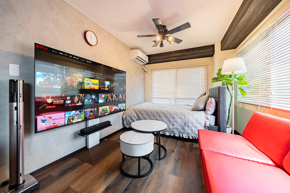 A living area features a large 4K television mounted on the wall, displaying streaming service options. A spacious bed is positioned nearby, with sunlight streaming through partially covered windows. Two circular tables in front of a red sofa create a cozy gathering space.