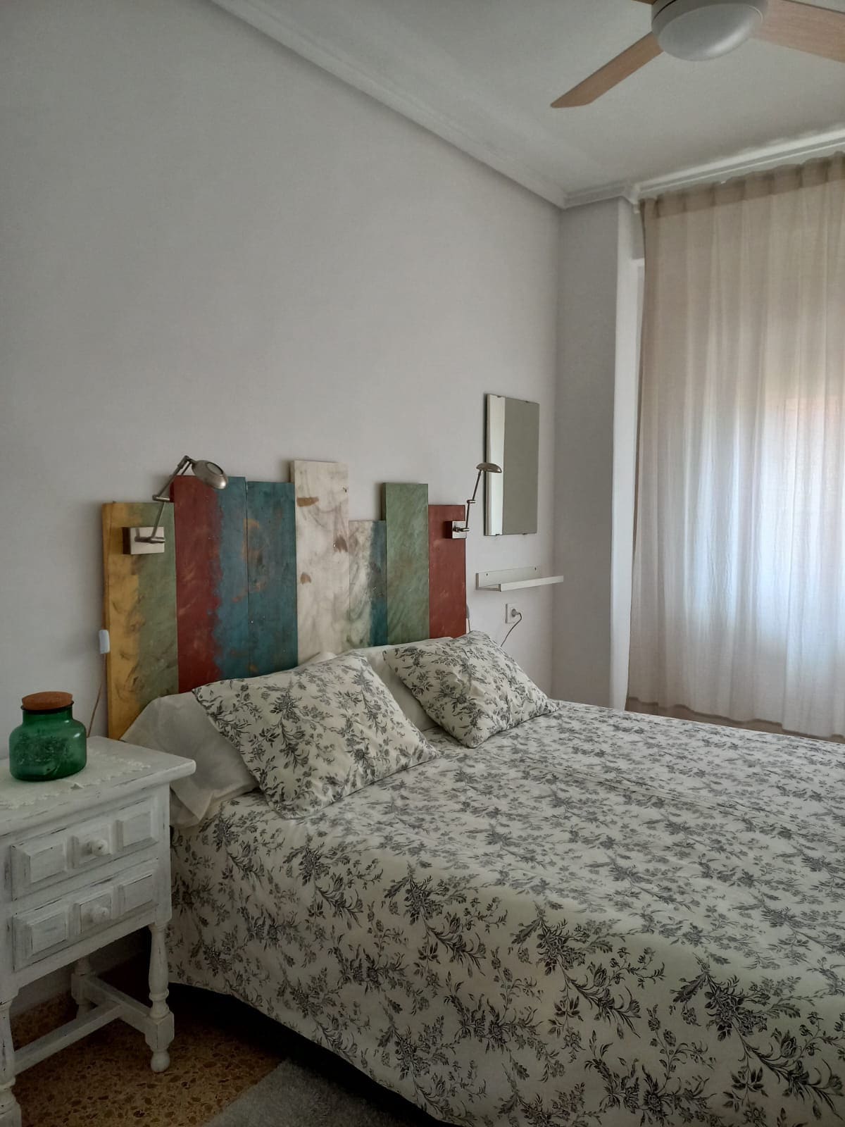 A bright bedroom features a spacious bed adorned with a floral-patterned duvet and pillows. A colorful wooden headboard adds character, while natural light filters through sheer curtains. A small bedside table with a green glass lamp is positioned beside the bed, enhancing the room's functionality.