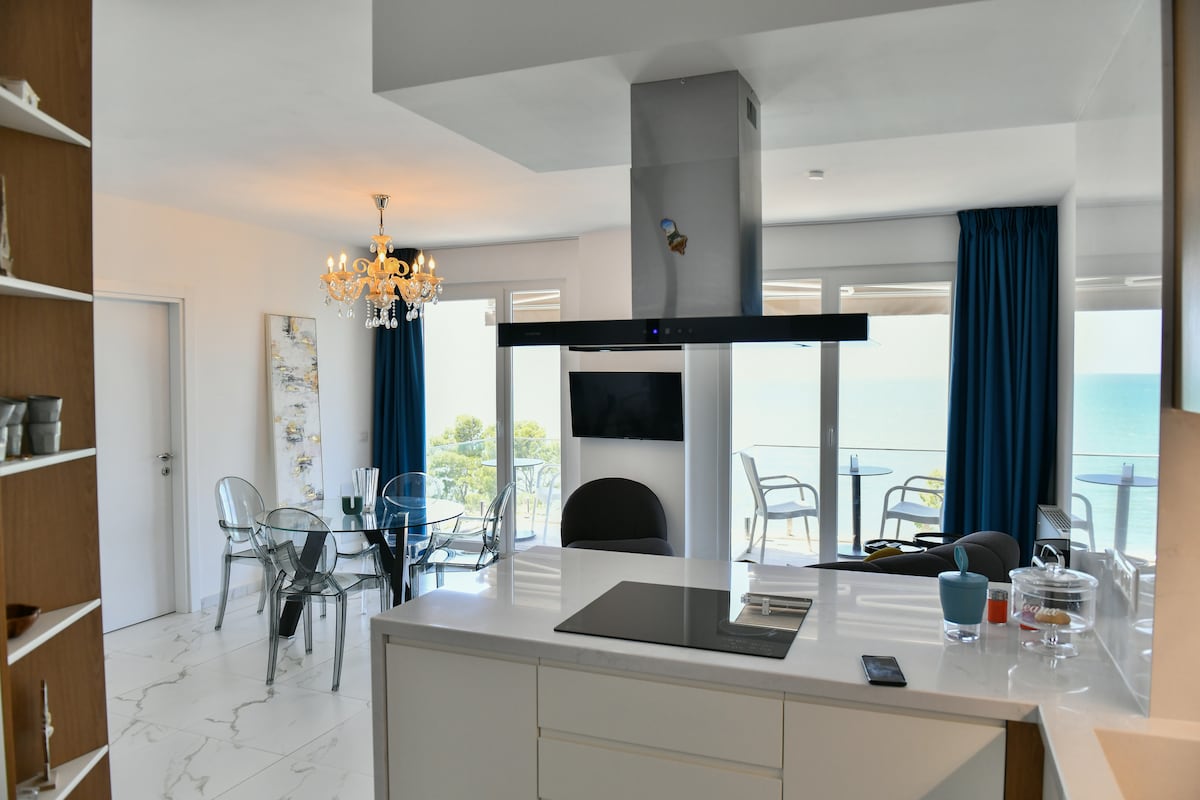 A modern kitchen with a sleek island is visible, featuring a stovetop and minimalist design elements. Adjacent to the kitchen, a dining area with transparent chairs overlooks the sea through large windows, with curtains framing the natural light. A chandelier adds elegance to the space.