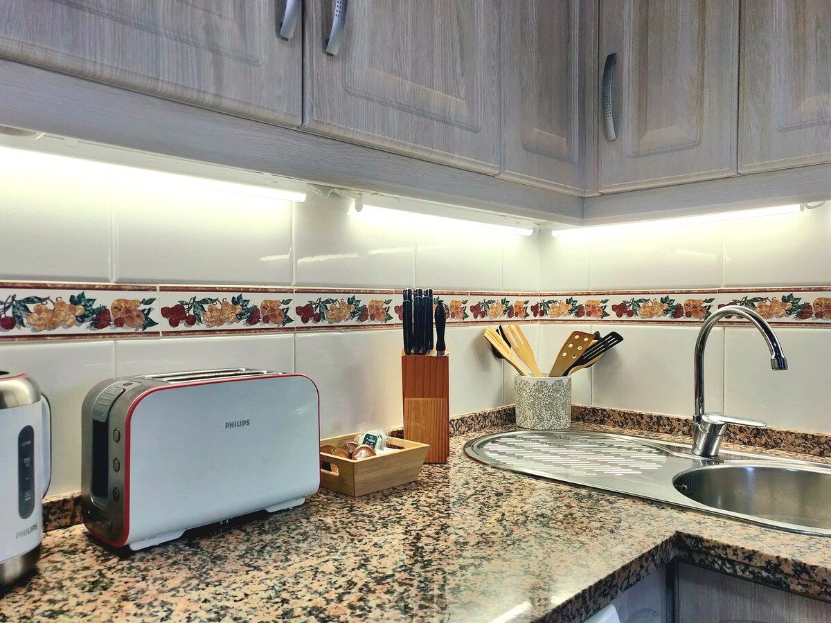 A modern kitchen corner is highlighted by a granite countertop. Essential appliances, including a silver toaster, are featured alongside a neat arrangement of utensils and knives in a wooden holder. Under-cabinet lighting adds clarity to this functional space.