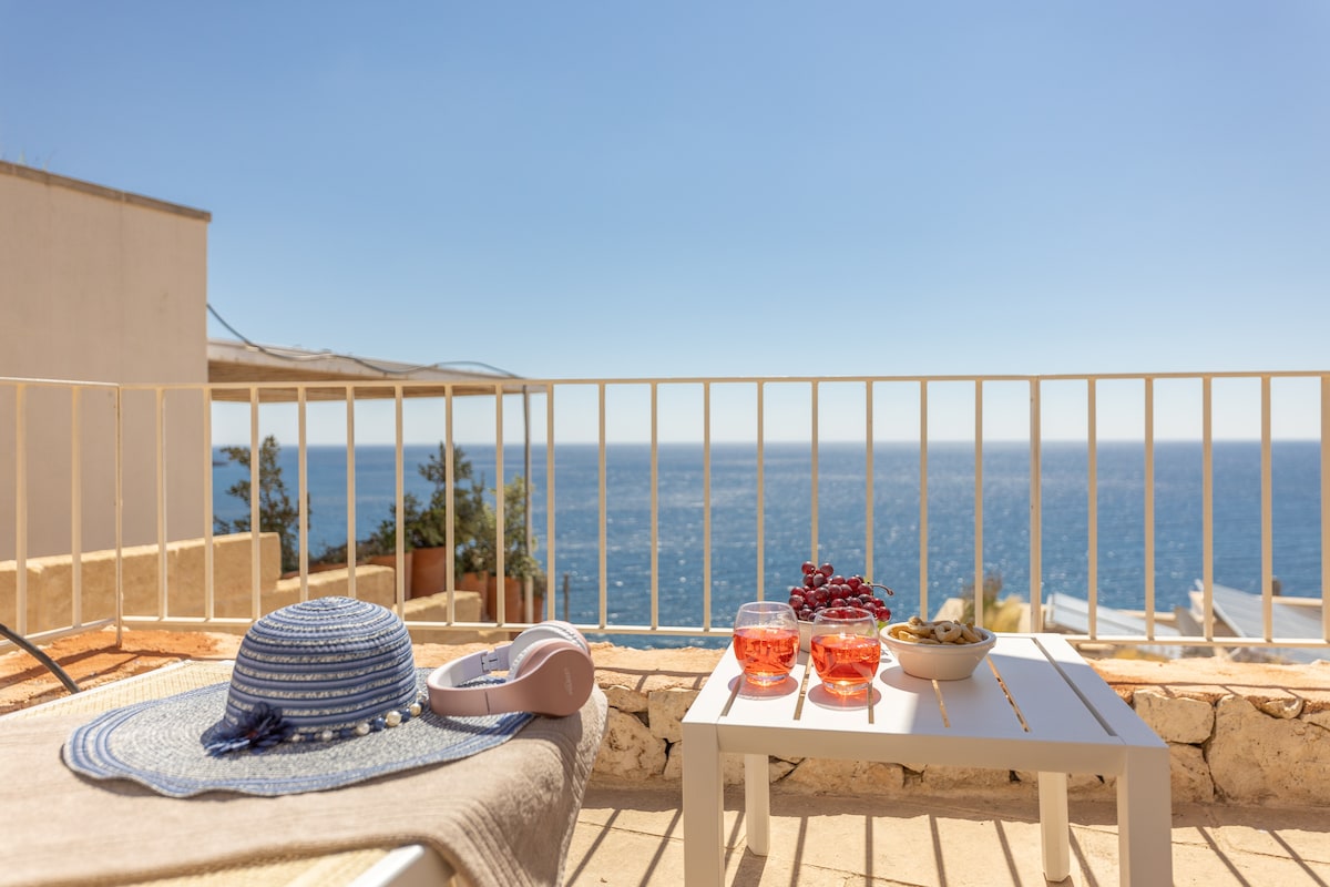 A sunlit terrace offers a view of the expansive sea, complemented by a small white table featuring glasses filled with colorful beverages and a bowl of olives. A wide-brimmed hat and headphones are placed on a lounge chair, inviting relaxation.