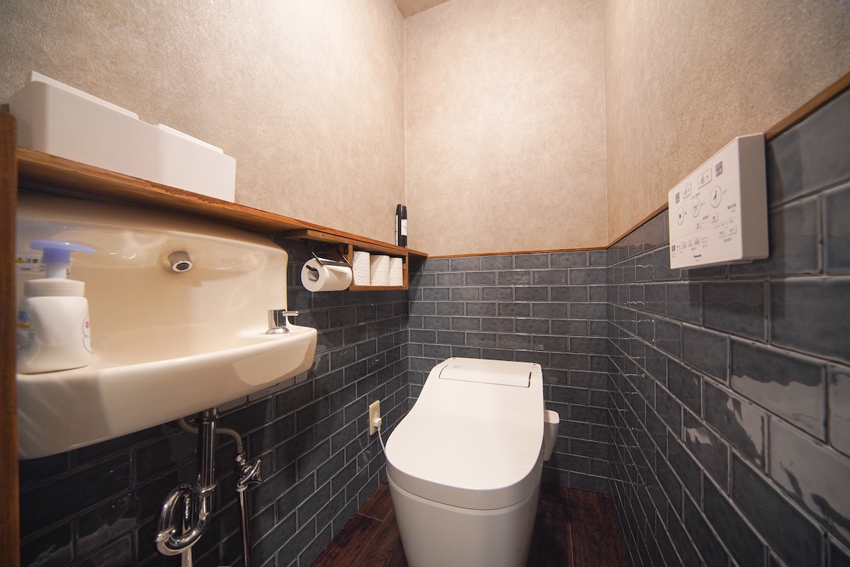A well-designed restroom features a modern toilet and a small sink with a minimalist faucet. Shelves above the sink hold toiletries and rolls of tissue. The walls are tiled in dark blue with a subtle textured finish, enhancing the overall sleek look.