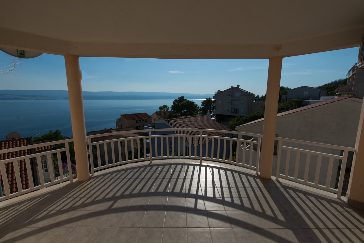 A large balcony offers expansive views of the sea and surrounding landscape. The floor is tiled, and sunlight casts shadows across the surface. The railings are white, enclosing the space and providing a sense of openness to the tranquil scenery.