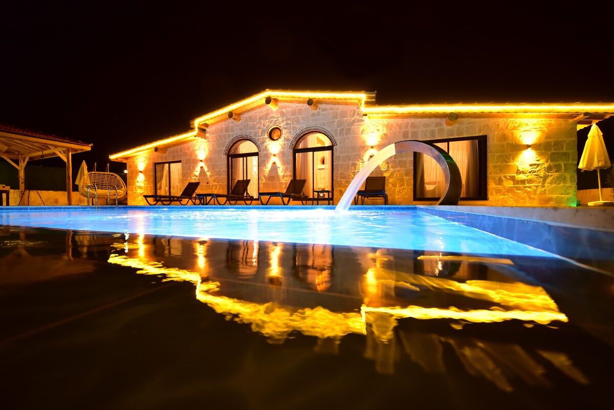 The villa features a stone façade illuminated by soft lighting at night, with a private pool reflecting the warm glow. Lounge chairs surround the pool, and a gentle water feature adds to the serene ambiance.