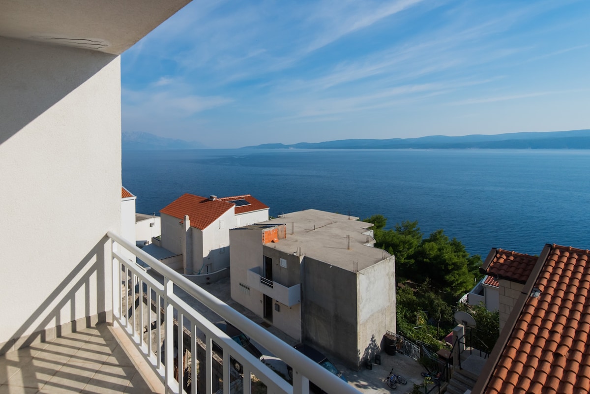 The image captures a balcony with white railings overlooking a serene sea view. The calm blue water reflects the sky, and buildings with terracotta roofs are visible nearby. Green foliage adds a touch of nature, enhancing the coastal setting.