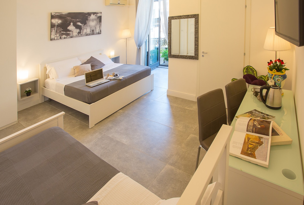 The spacious bedroom features a matrimonial bed and an additional single bed. A work desk is positioned near a wall-mounted television. Natural light is allowed in through a nearby balcony door, creating a bright and functional atmosphere. A mirror and decorative items complement the modern decor.