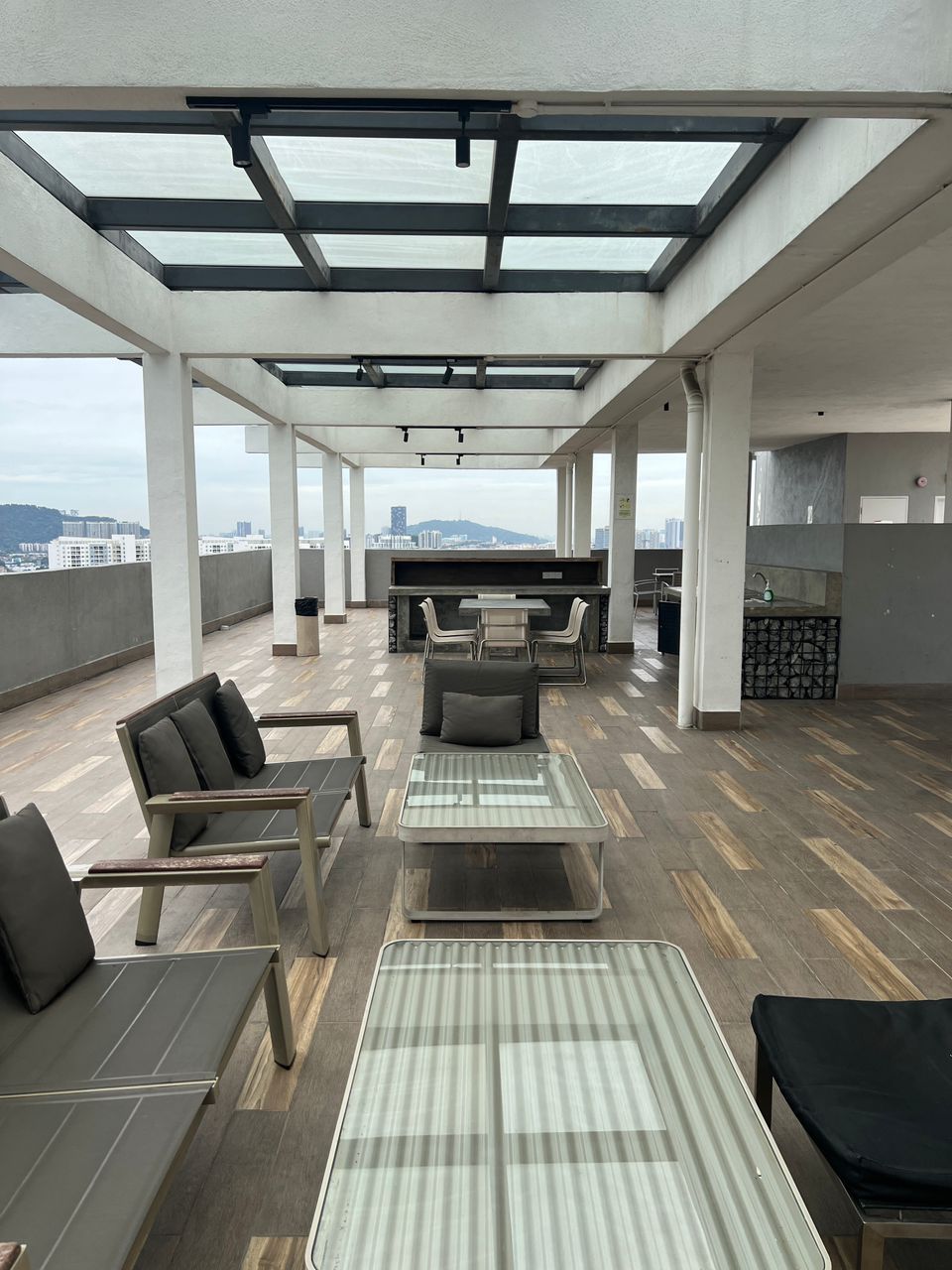 A spacious rooftop area is shown, featuring a combination of glass-topped tables and comfortable seating. Natural light floods the space through the overhead skylights, illuminating the wooden floor. A distant city skyline is visible beyond the railing, enhancing the open atmosphere.