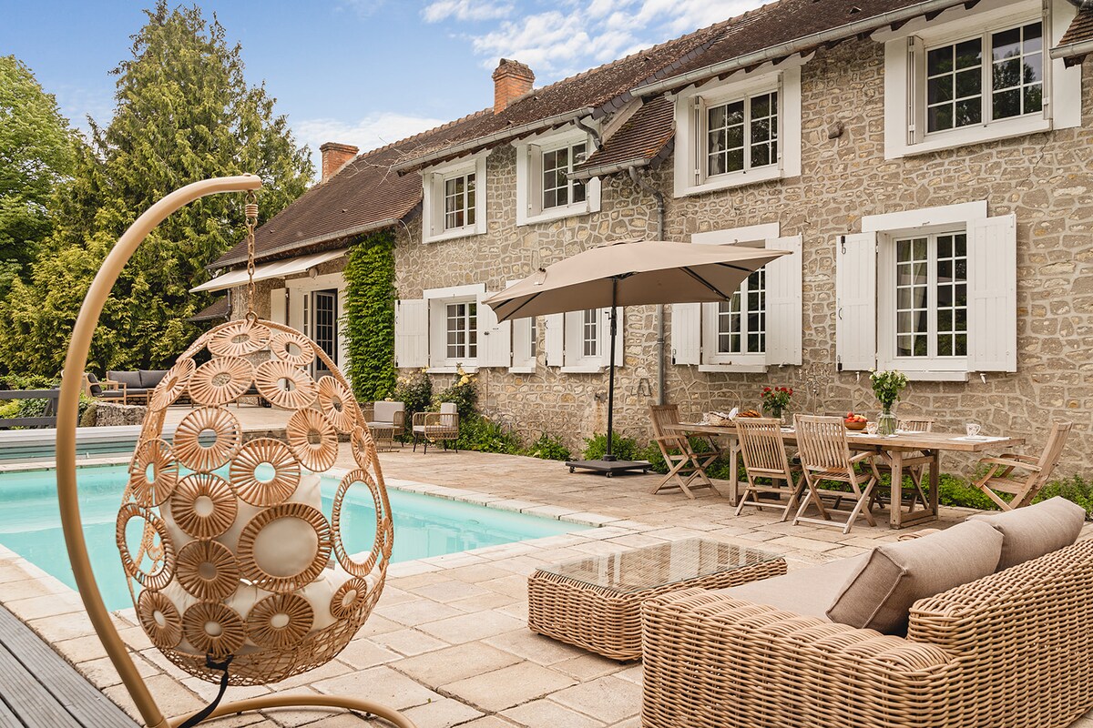 A charming stone house with white shutters is depicted alongside a serene pool area. Woven lounge chairs and a round swinging seat are positioned near the pool, offering a relaxed outdoor setting. A large umbrella provides shade over the patio furniture, inviting guests to enjoy the space.