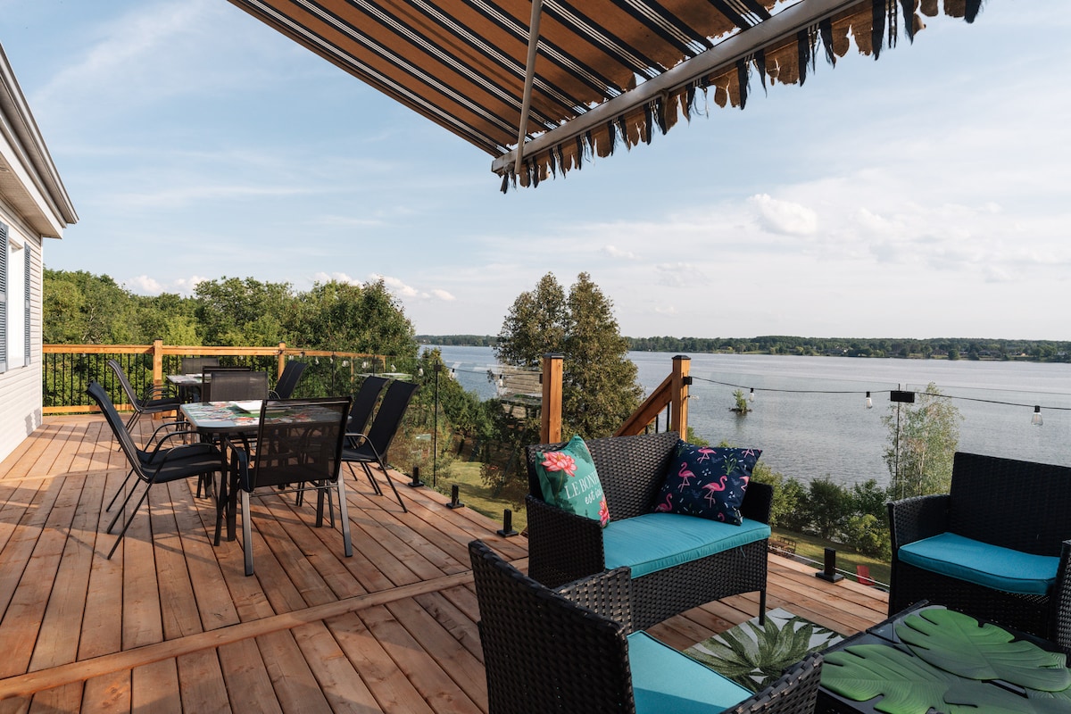 A spacious wooden deck features comfortable seating arrangements, including a table and chairs for dining al fresco. A retractable awning provides shade, and views of the water and surrounding nature are visible in the background.