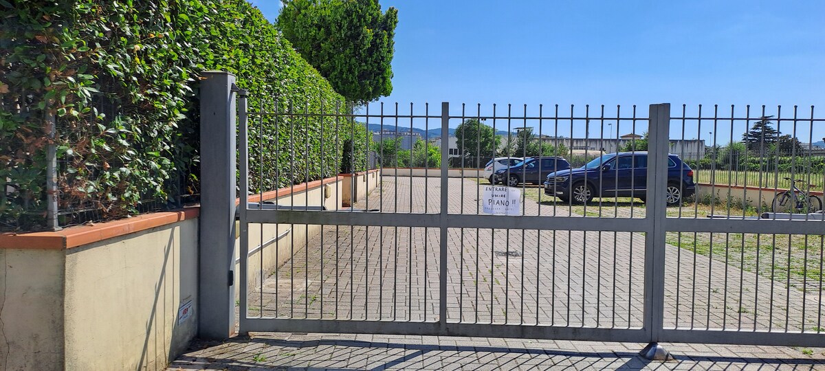 A gated entrance is shown, featuring a tall, gray metal gate supported by concrete pillars. A spacious parking area is visible behind the gate, with several parked cars and greenery lining the perimeter. A clear blue sky enhances the scene.