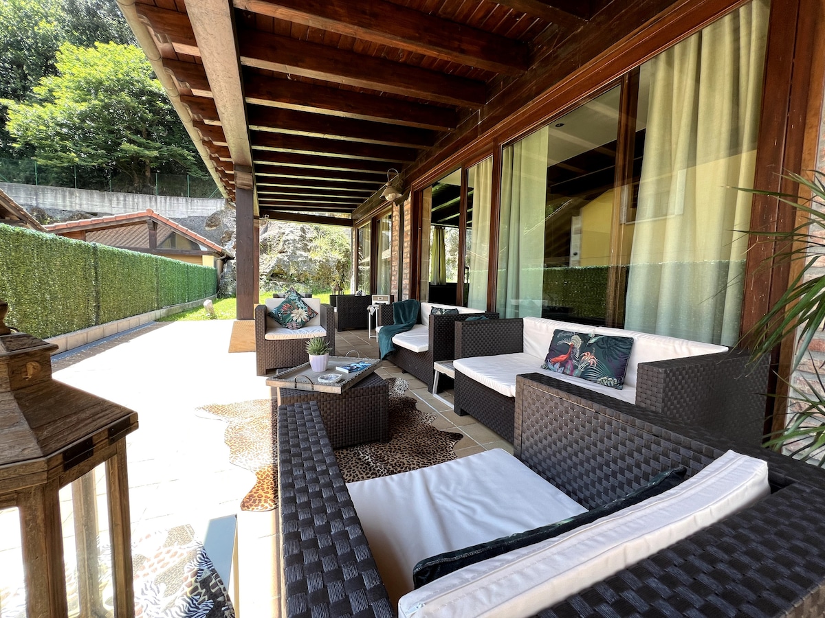 An outdoor seating area is displayed under a wooden pergola, featuring dark wicker furniture and soft cushions. Natural light fills the space through large windows, while potted plants add a touch of greenery. The surrounding garden is visible in the background, enhancing the relaxed ambiance.