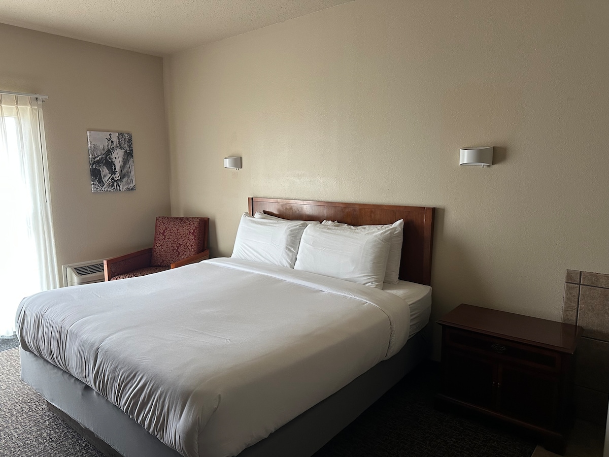 A spacious bedroom features a large bed with white linens. Soft lighting is provided by wall-mounted sconces. A seating area includes an upholstered chair next to a small side table, while a sliding glass door offers natural light.