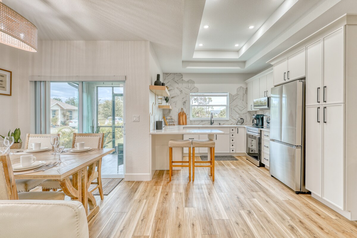 An open-plan kitchen and dining area is illuminated by natural light from large windows. A six-seat dining table is placed next to a counter with bar stools. Sleek cabinetry and stainless-steel appliances create a modern look, while neutral tones and wood flooring enhance the inviting atmosphere.