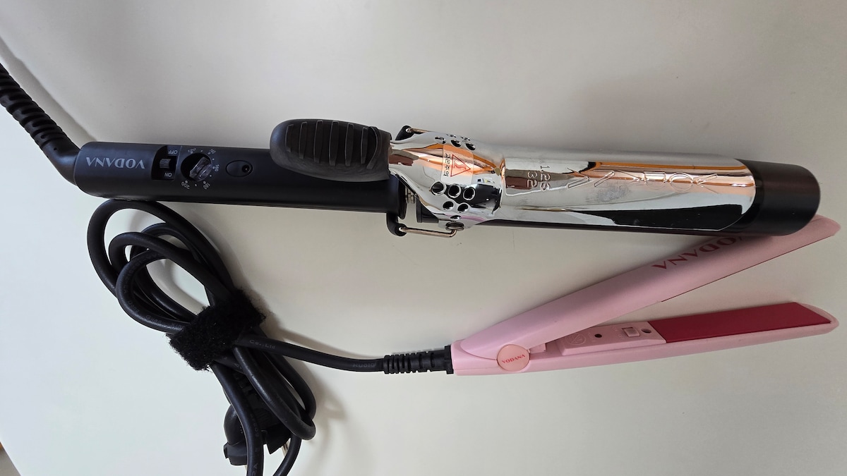 A hair styling tool is displayed, featuring a silver and black curling iron alongside a pink flat iron. Both tools have power cords that are neatly arranged. The curling iron includes a control panel for temperature adjustments.