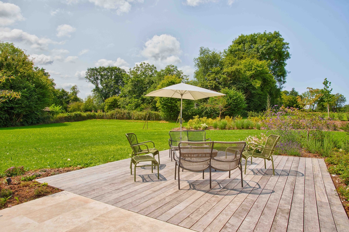 A wooden deck features a set of six metal chairs and a round table, arranged under a large umbrella. Surrounding greenery and flower beds are visible, contributing to a peaceful outdoor setting. A spacious lawn extends beyond the deck, inviting relaxation.