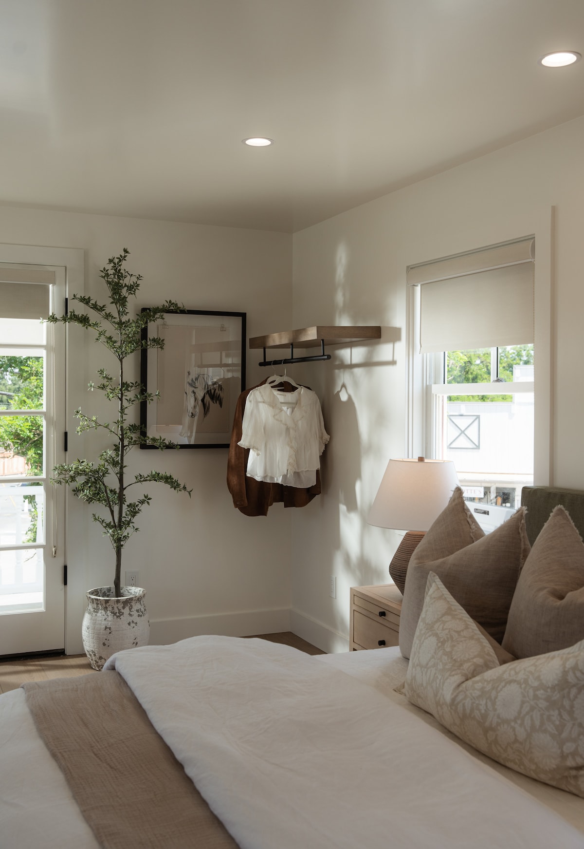 A bright and airy bedroom is presented with large windows allowing natural light to enter. A cozy bed is adorned with soft pillows, while a decorative plant adds a touch of greenery. Clothing is neatly displayed on a wall-mounted rack, contributing to the tidy atmosphere.