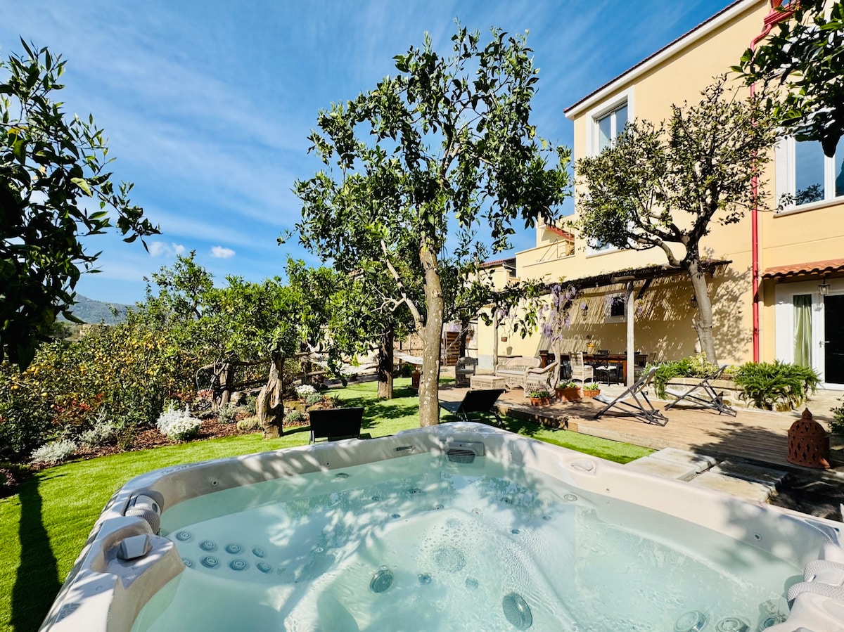 A relaxing outdoor area features a hot tub situated amidst lush greenery, including small trees laden with fruit. The surrounding garden showcases vibrant plants and well-maintained grass, while a portion of the villa is visible in the background, adding to the serene ambiance.