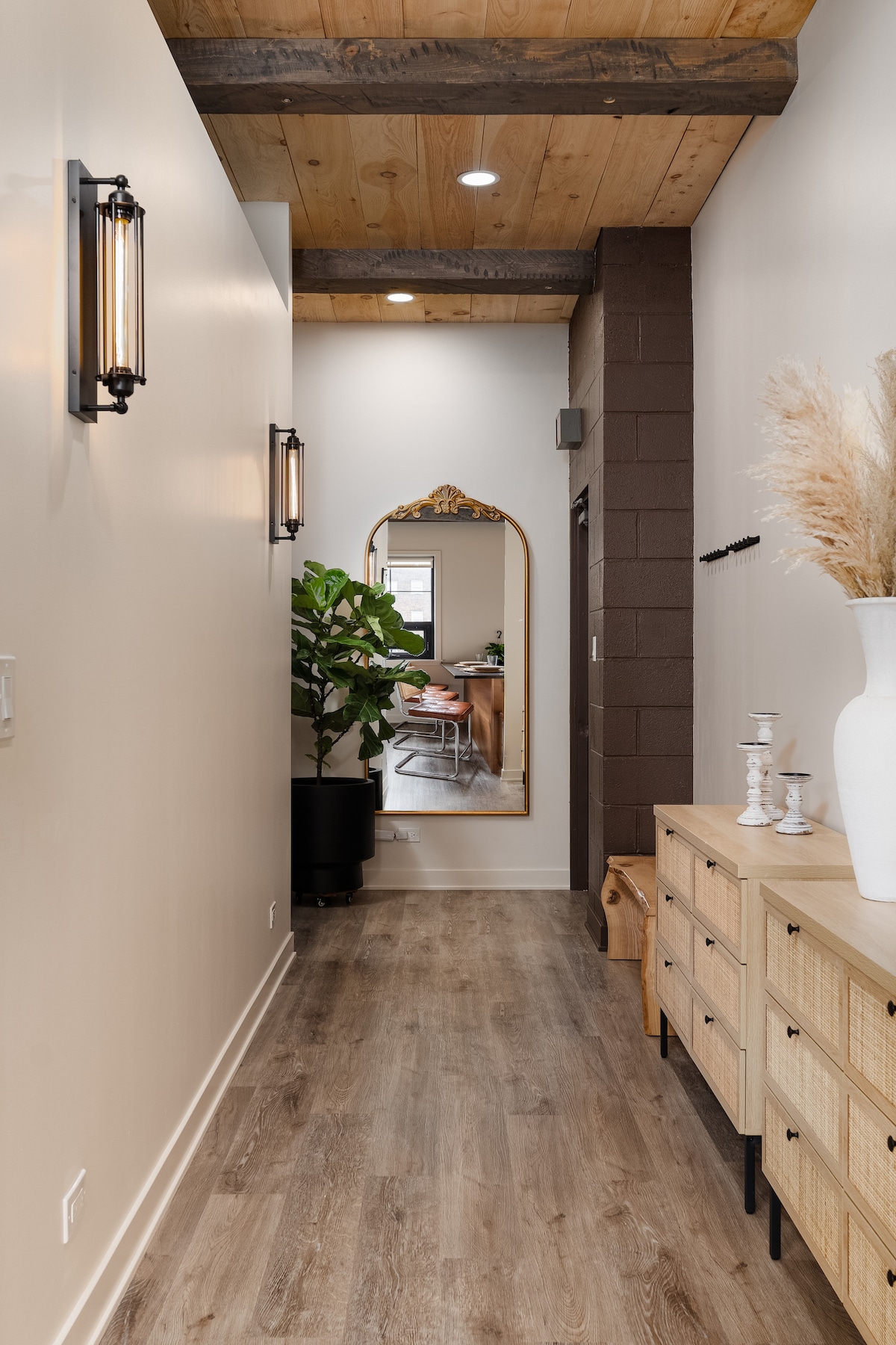 A narrow hallway features warm wood beams overhead and stylish wall sconces. A large mirror reflects the space, while a tall green plant adds a touch of nature. A console with woven drawers and decorative items complements the inviting atmosphere.
