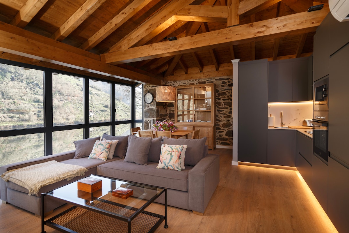 A spacious living area features a large gray sofa with decorative pillows and a wooden coffee table. Bright windows allow natural light to fill the room, offering views of the surrounding landscape. The modern kitchen is equipped with sleek cabinetry, and a dining table is placed nearby.