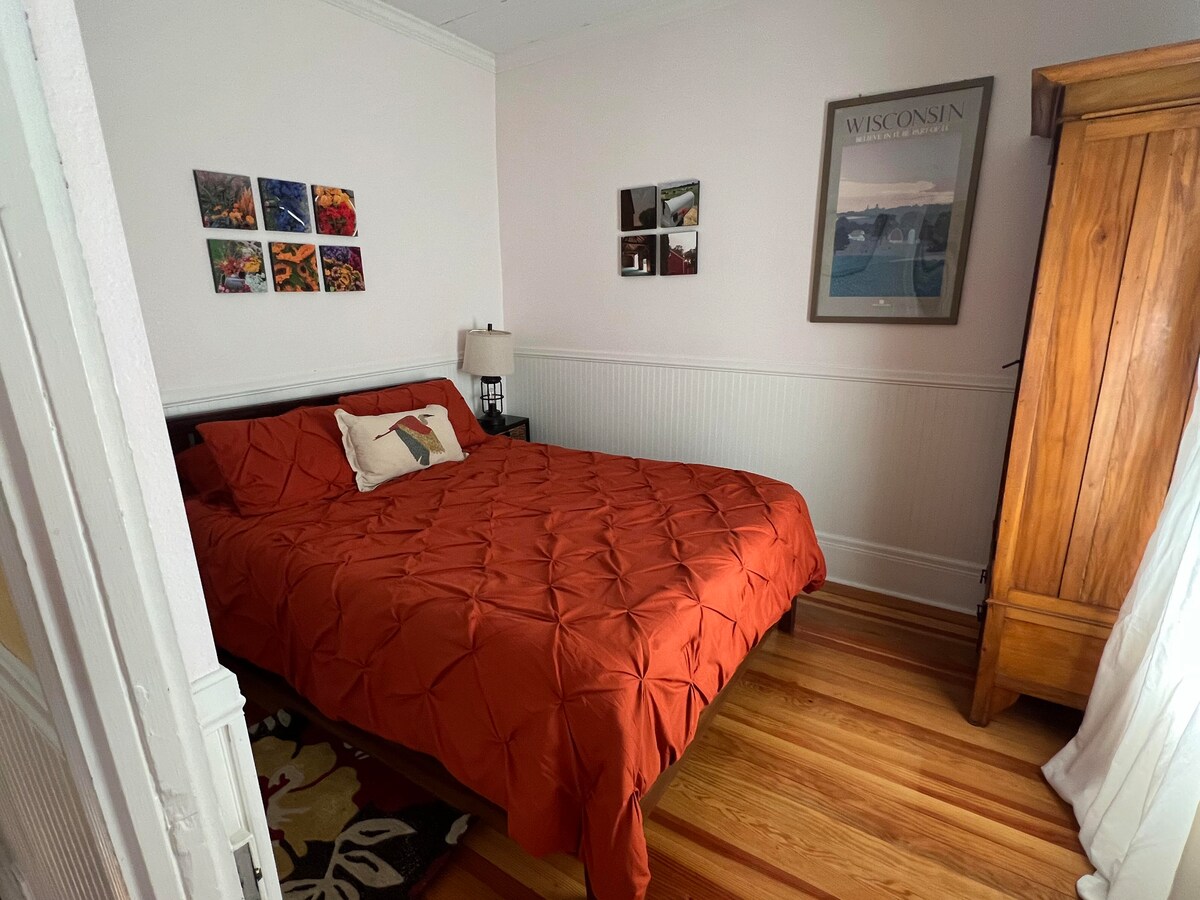 Hay una cama tamaño queen en el dormitorio delantero, con un armario antiguo y mesitas de noche con lámparas a cada lado.