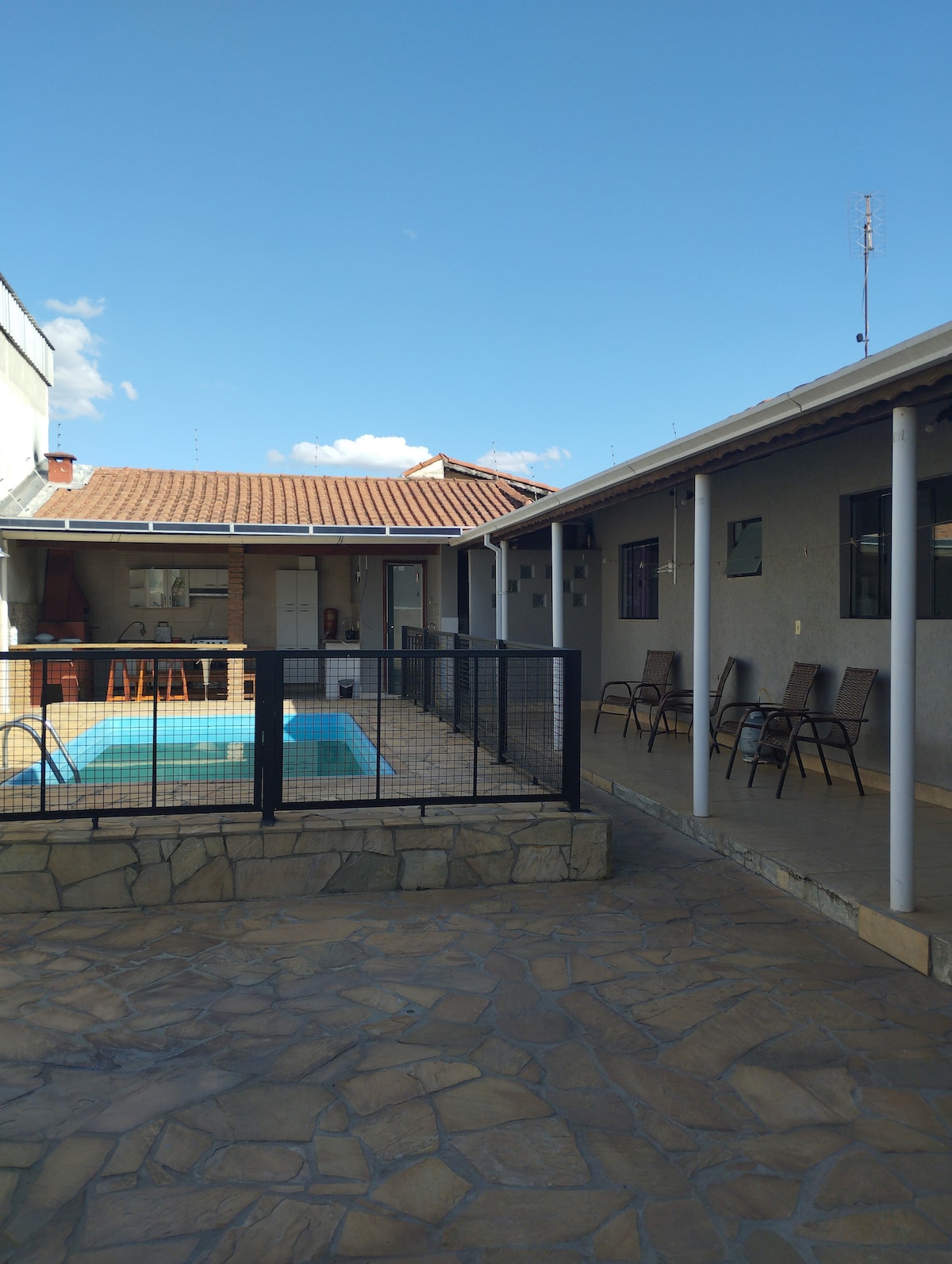 The backyard features a swimming pool surrounded by a safety fence, with a spacious patio area and a stone pathway. Several chairs are positioned around the pool, and the interior space is visible through glass doors, offering an inviting view of the main living area.
