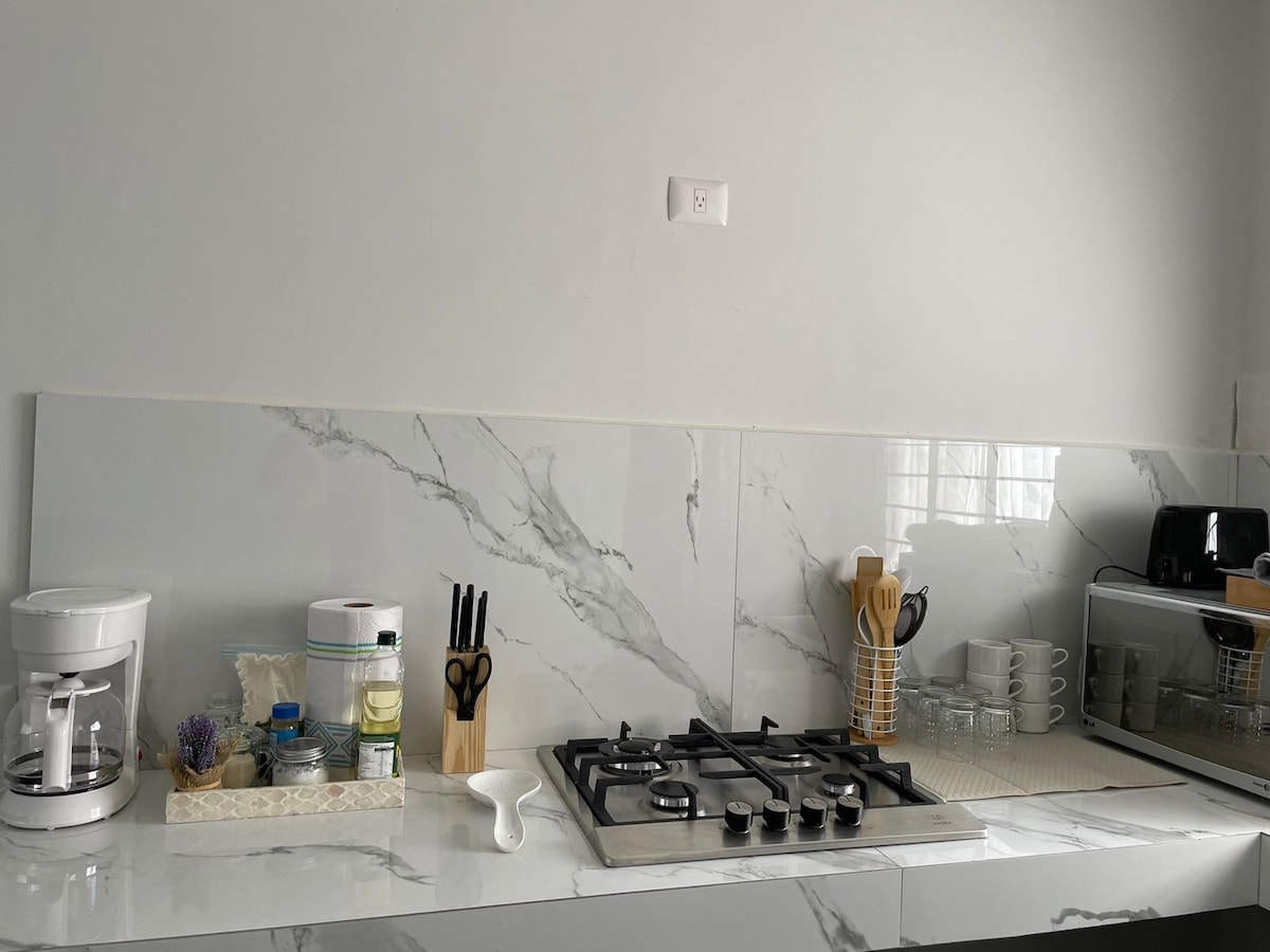 A modern kitchen area features a marble countertop, equipped with various kitchen appliances including a coffee maker, blender, and toaster. An organized array of utensils and dishes is visible, alongside a gas stove with multiple burners. Natural light brightens the space.