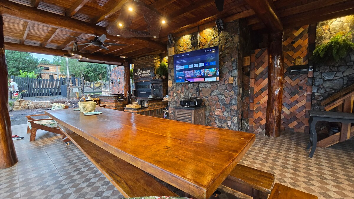 A large wooden dining table is positioned in a spacious outdoor area. Stone walls and wooden beams provide a natural aesthetic, while a flat-screen television is mounted on one wall. Lights are arranged to create an inviting ambiance, suitable for gatherings.
