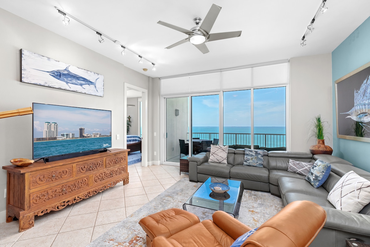 A bright living area features a modern sectional sofa and a large television. Floor-to-ceiling windows offer unobstructed views of the ocean. Art decorates the walls, and natural light fills the space through the glass doors leading to the balcony.