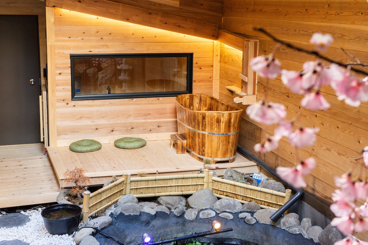 A serene outdoor space features a wooden hot tub surrounded by natural stone and polished wood surfaces, with two green seating cushions nearby. Cherry blossom branches frame the image, adding a touch of nature to the peaceful environment.