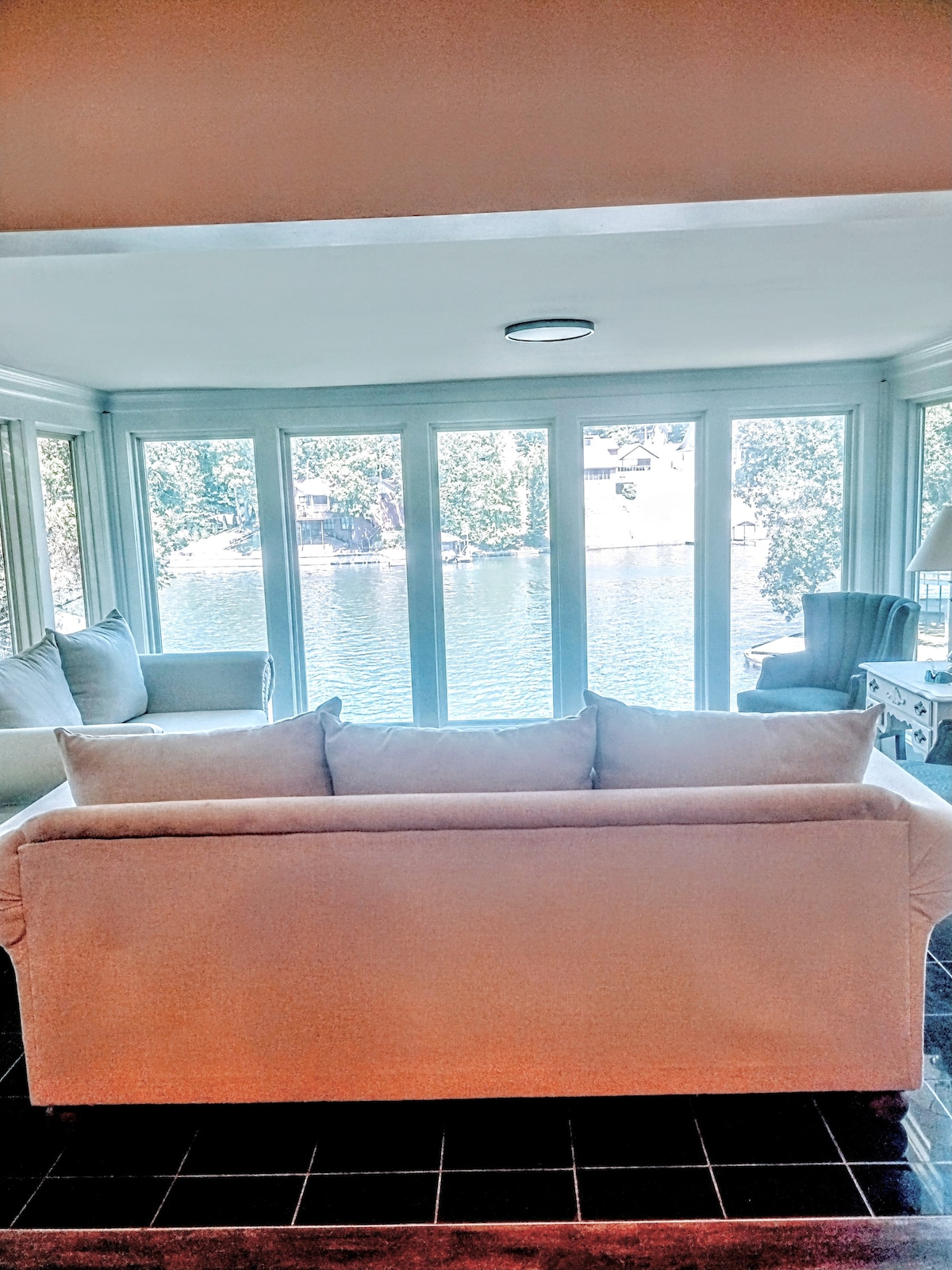 A comfortable living area is showcased with a neutral-colored sofa facing large glass doors that offer a direct view of the tranquil lake. The space includes additional seating and natural light flooding in from the windows, enhancing the serene atmosphere.