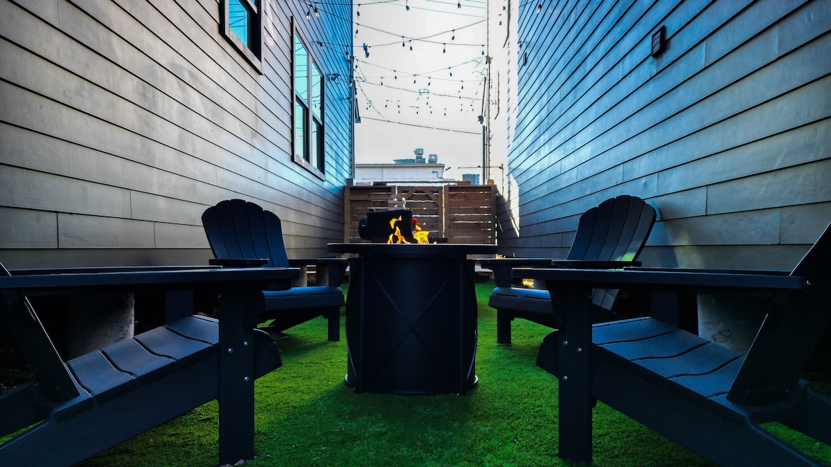 An outdoor patio area features several adirondack chairs arranged around a central fire pit. Green artificial grass covers the ground, and string lights are hung above, creating an inviting space for gatherings.